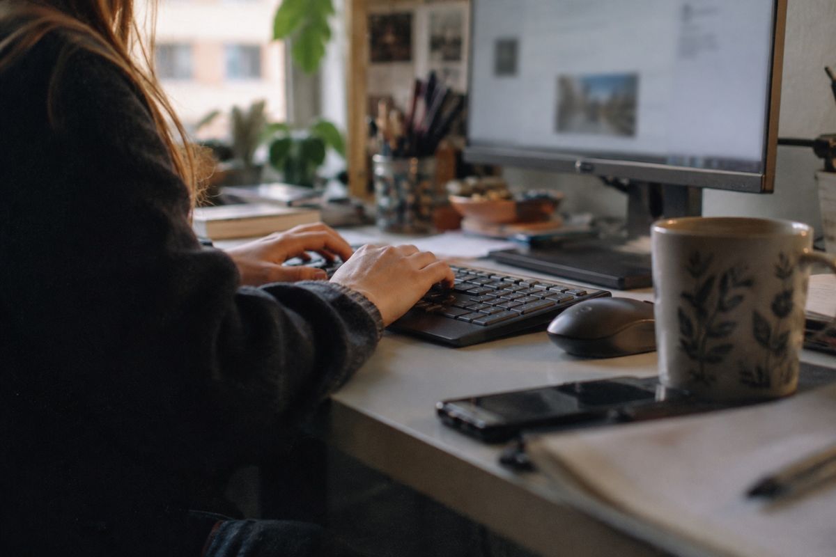 Eine Frau tippt am Schreibtisch auf einer Tastatur vor einem großen Monitor. Daneben liegen eine Maus und ein Smartphone, im Vordergrund steht eine Tasse mit Blättermuster.